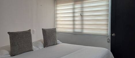 A bedroom with a bed, two pillows, a window with blinds, and a door.