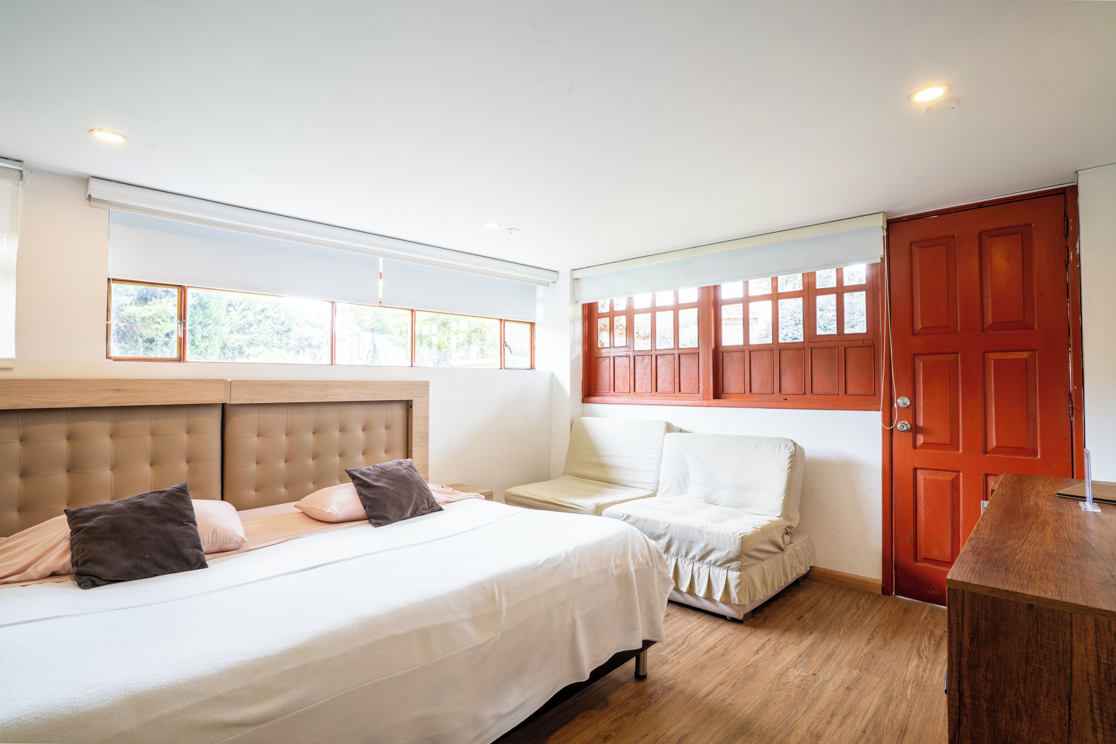 A bedroom with a large bed, a chair, a wooden desk, and a red door.