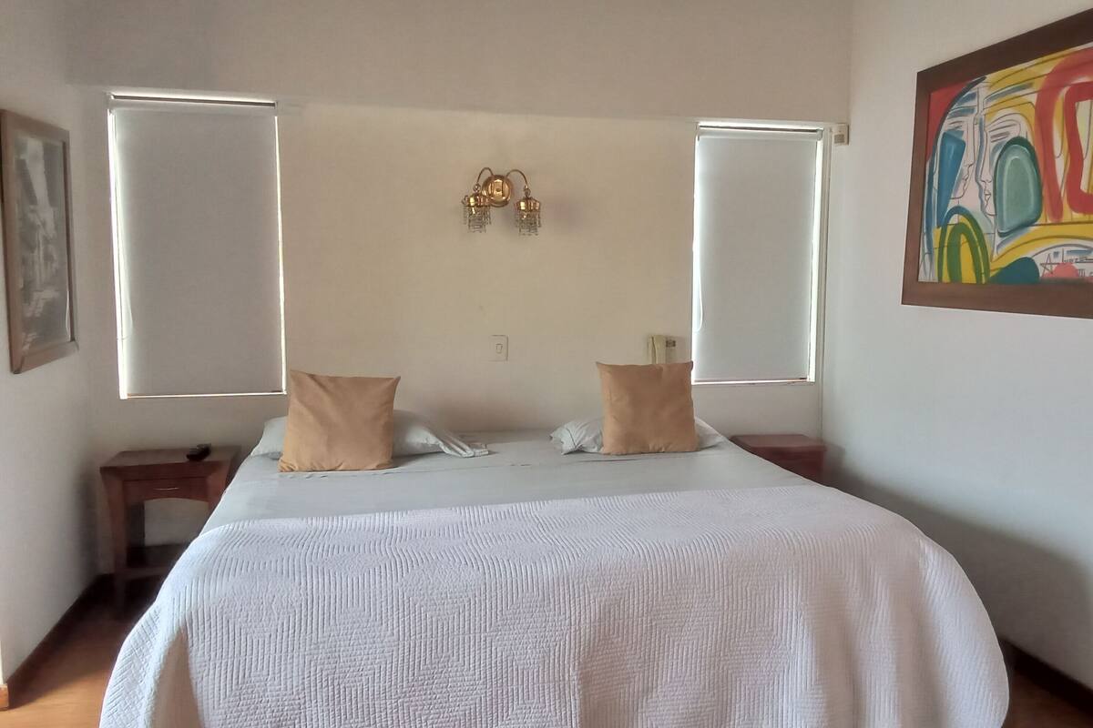 A bedroom with a bed, two pillows, a nightstand, a wall clock, a window with blinds, and a painting.