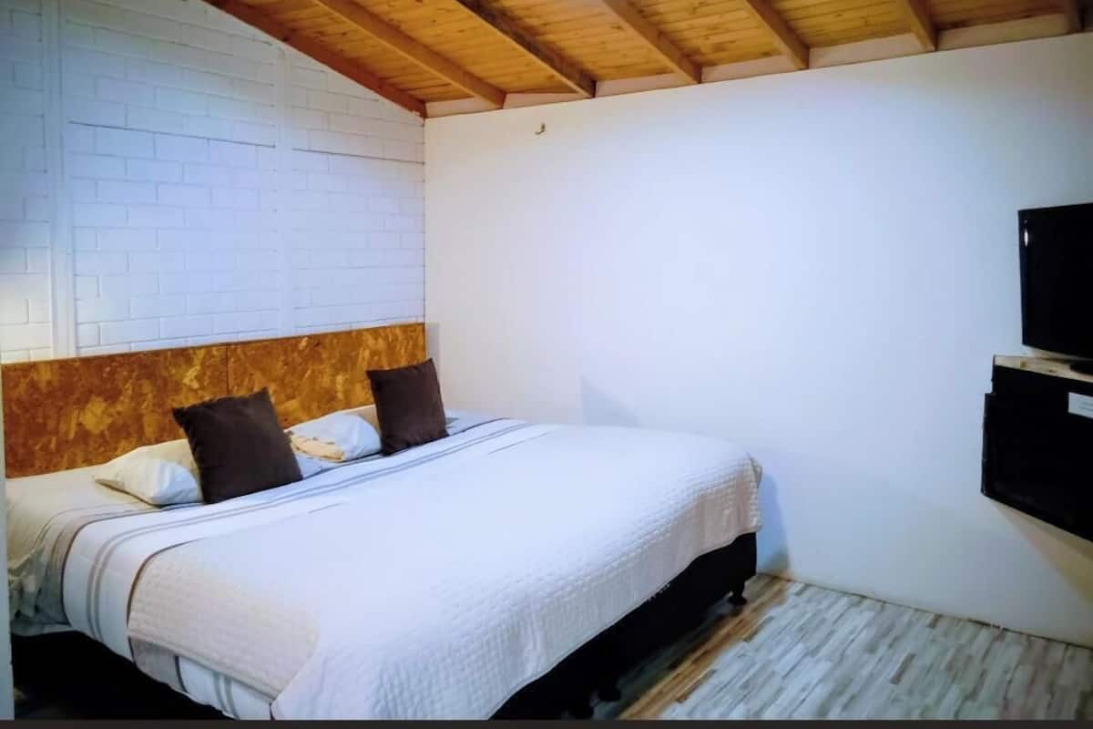 A bedroom with a wooden ceiling, a white brick wall, a bed with a white bedspread, and a mounted television.