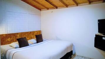 A bedroom with a wooden ceiling, a white brick wall, a bed with a white bedspread, and a mounted television.