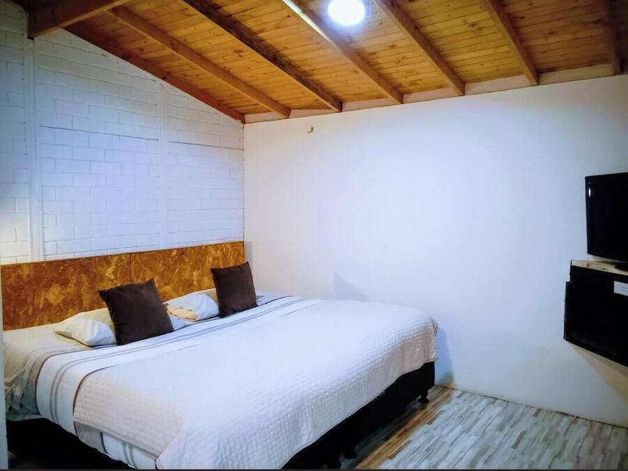 A bedroom with a wooden ceiling, a white brick wall, a bed with a white bedspread, and a mounted television.