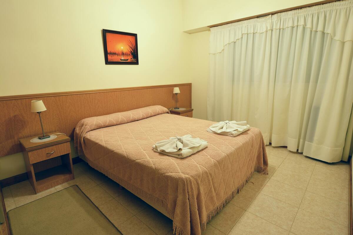 Un lit bien fait, recouvert d’une couverture beige, deux lampes de chevet, une table de nuit, un tableau encadré au mur et une fenêtre avec des rideaux blancs.