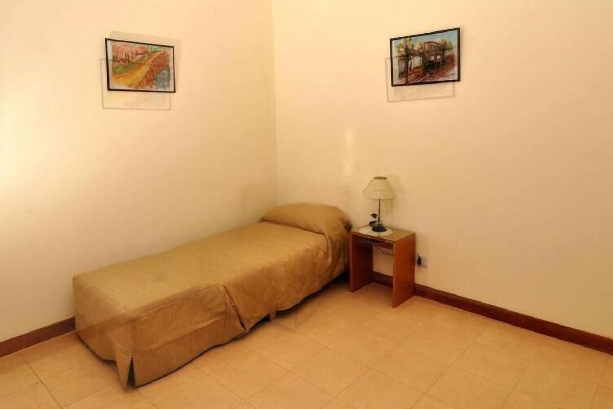 Une petite chambre avec un lit simple, une table de chevet en bois et une lampe, deux tableaux encadrés aux murs et un sol carrelé.