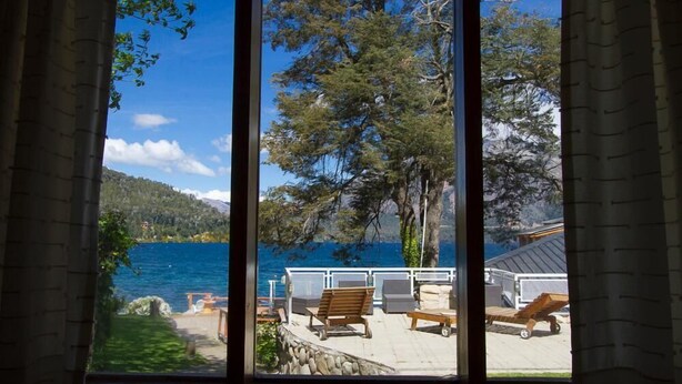 A view through a window of a terrace with outdoor furniture, a stone wall, and a body of water with trees and hills in the background. - Los Castaños (San Carlos de Bariloche)