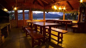 A rustic wooden dining area with benches and tables, large windows with mountain views, and a stone fireplace. - Los Castaños (San Carlos de Bariloche)