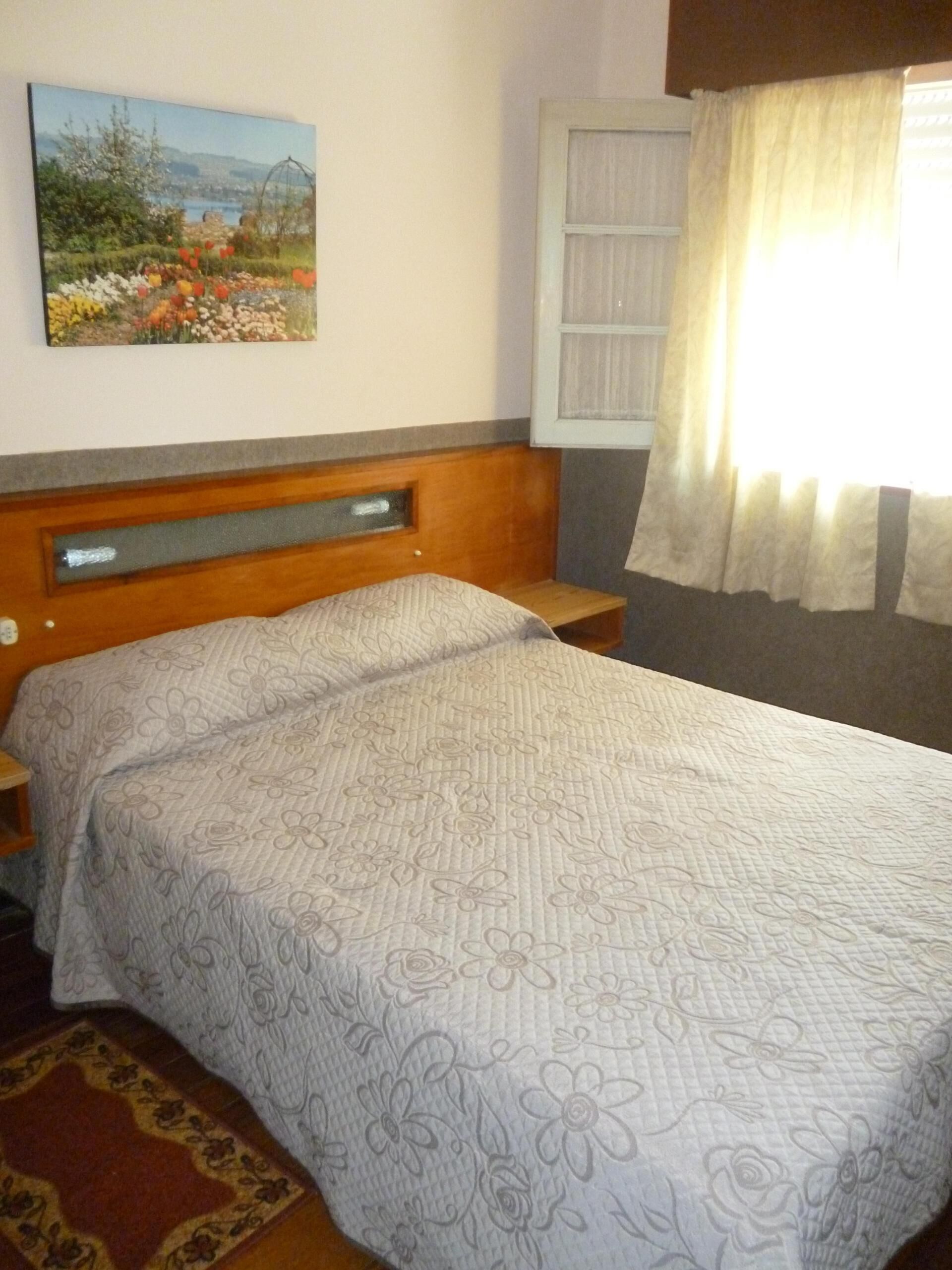 Een slaapkamer met een bed, nachtkastje, houten hoofdbord, raam met gordijnen en een schilderij aan de muur.