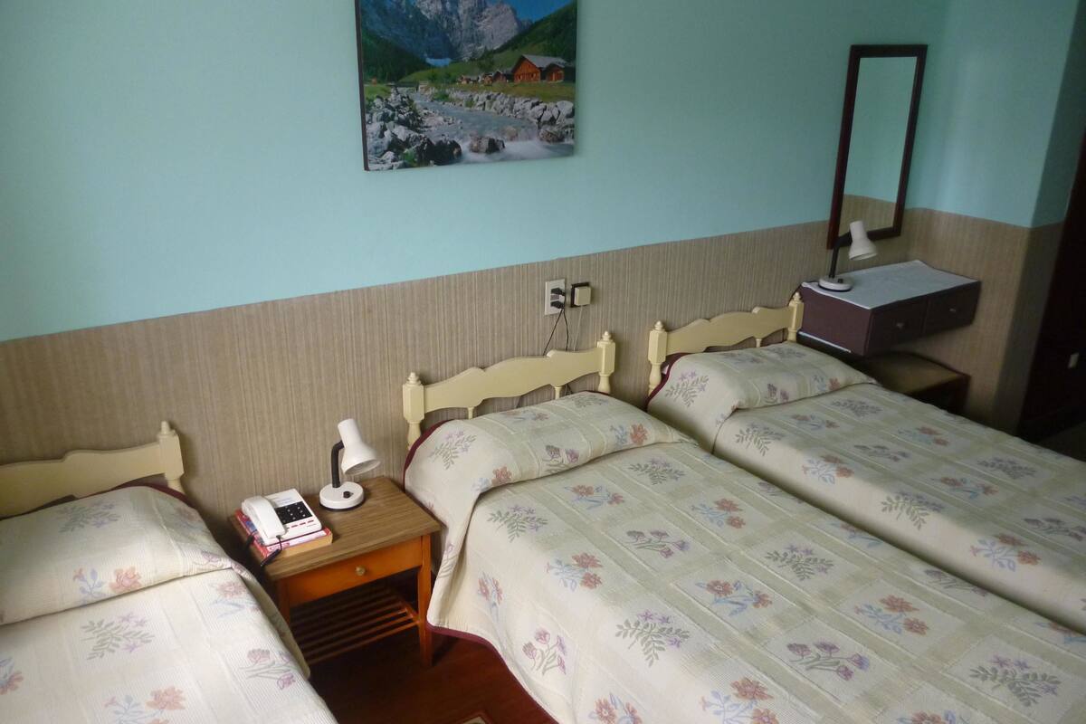 Ein Hotelzimmer mit zwei Einzelbetten, einem Nachttisch mit Lampe, einem kleinen Schreibtisch ebenfalls mit Lampe und einem an der Wand befestigten Spiegel.