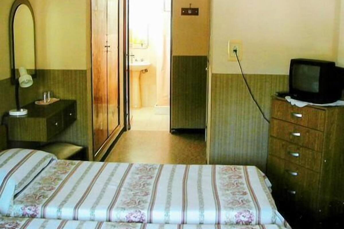 Ein Hotelzimmer mit einem Bett, einem Schreibtisch mit Lampe, einem Fernseher auf einer Kommode und einem durch eine offene Tür einsehbaren Badezimmer.
