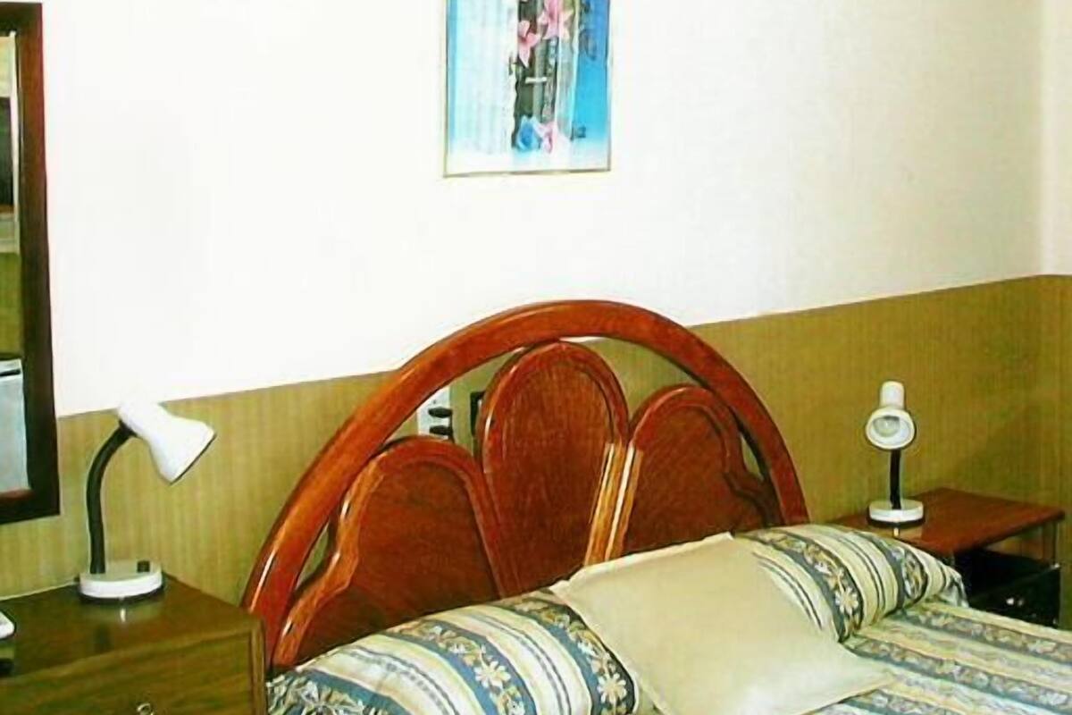 Ein Schlafzimmer mit Holz-Kopfteil, einem Bett mit gemusterten Betttextilien, einem Nachttisch mit Lampe und einem Bild an der Wand.
