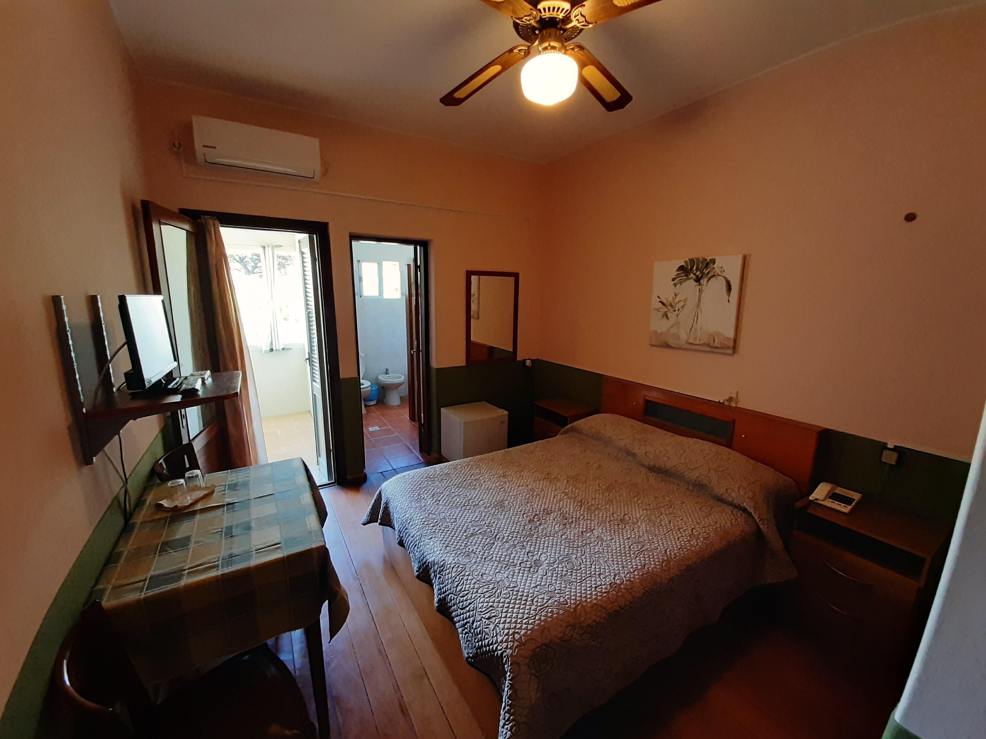 Un dormitorio con cama, escritorio con computadora, ventilador de techo y un cuadro en la pared.