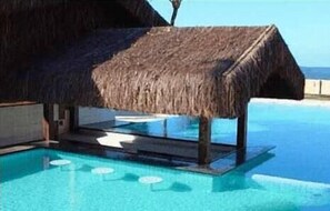 A poolside structure with a thatched roof, a clear blue pool, and a view of the ocean. - Bouganville Hotel (Beberibe)