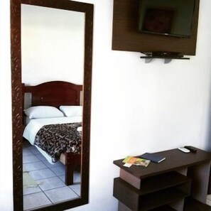 A hotel room with a bed, a desk with a chair, a mirror, and a television mounted on the wall. - Pousada Juliane (Penha)