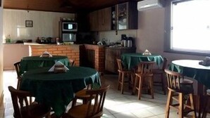 Un comedor con mesas cubiertas de manteles verdes, sillas de madera y una zona de cocina con microondas y refrigerador.