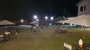 An outdoor patio area with tables and chairs under umbrellas, surrounded by a grassy lawn and a stone building. - Hostería ACA Tafí del Valle (Tafi del Valle)