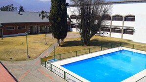 A swimming pool with a green fence, a building with arched windows, and a tree in the foreground. - Hostería ACA Tafí del Valle (Tafi del Valle)