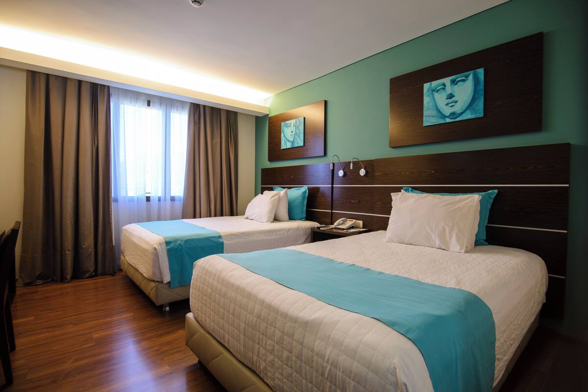 A hotel room with two beds, a wooden floor, a large window with curtains, and artwork on the walls.