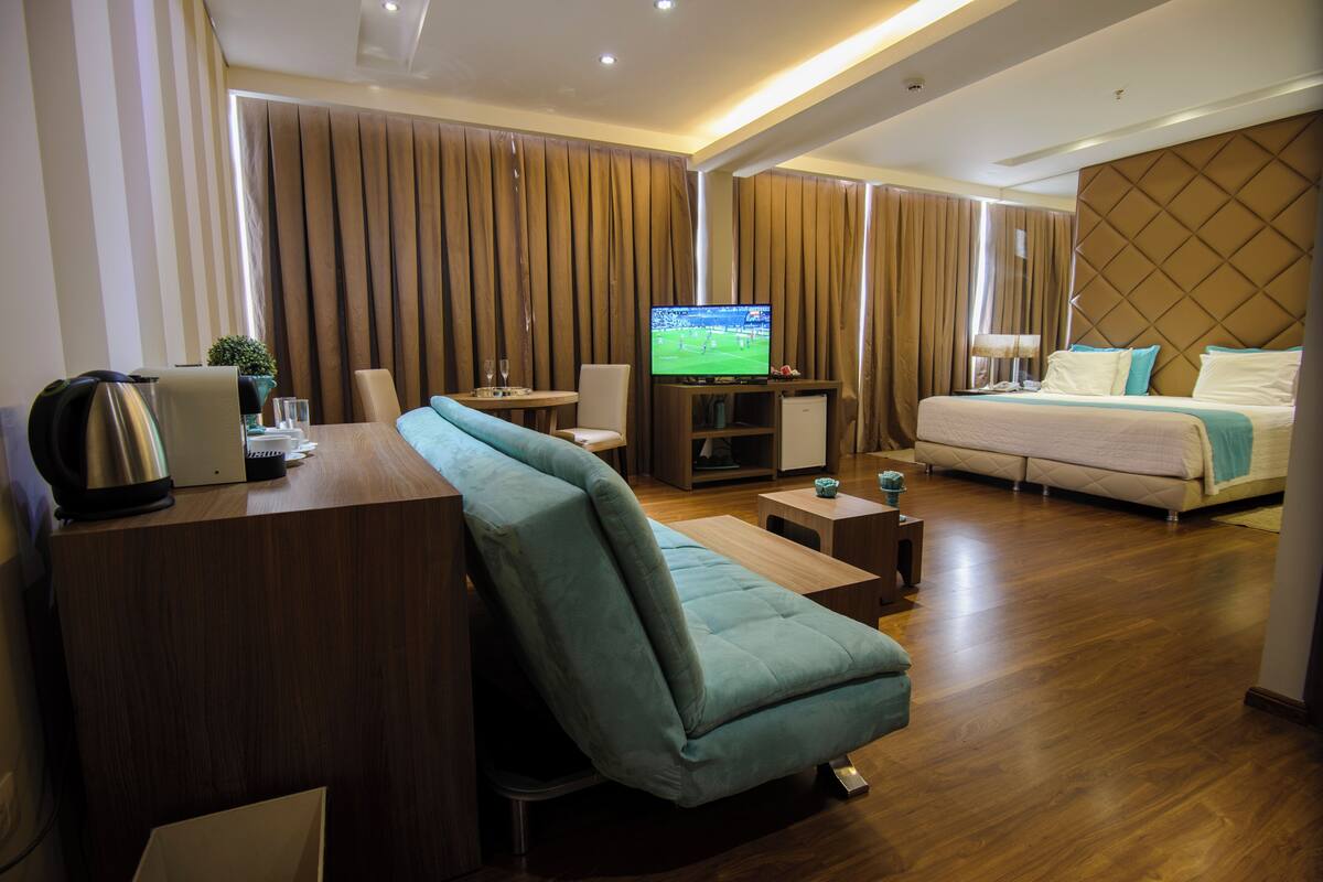 A modern hotel room with a flat-screen TV, a sofa, a bed, a desk with a kettle, and a small table.