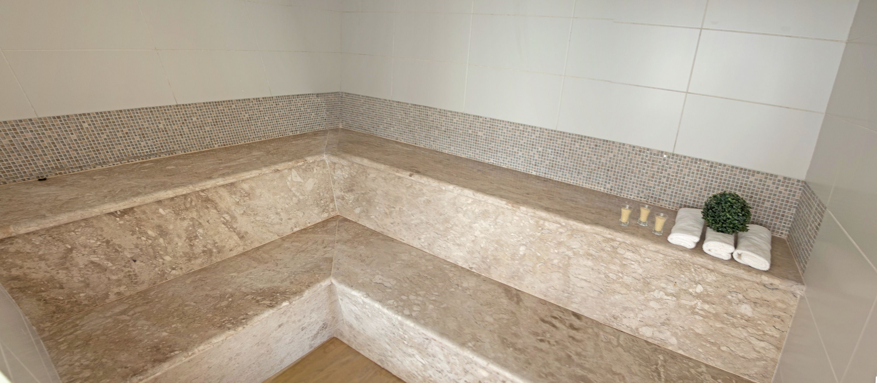 A corner bathtub with a tiled mosaic border, a marble countertop, and a small plant and towels on the side.