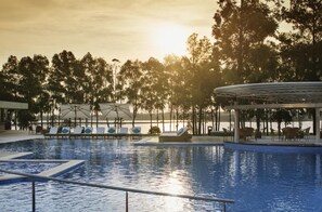 A swimming pool with lounge chairs and umbrellas, surrounded by trees and a view of the sunset. - Awa Resort Hotel (Encarnacion)