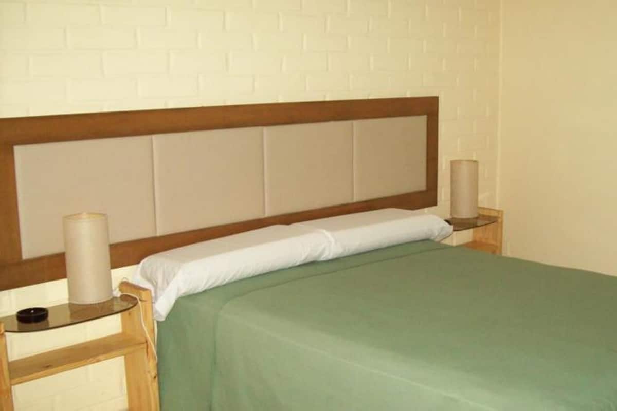 A single bed with a wooden headboard and green bedding, two bedside tables with lamps, and a white brick-patterned wall.