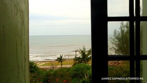A view through a window of a coastal area with a beach, palm trees, and a clear sky. - Recanto da Prainha (Torres)