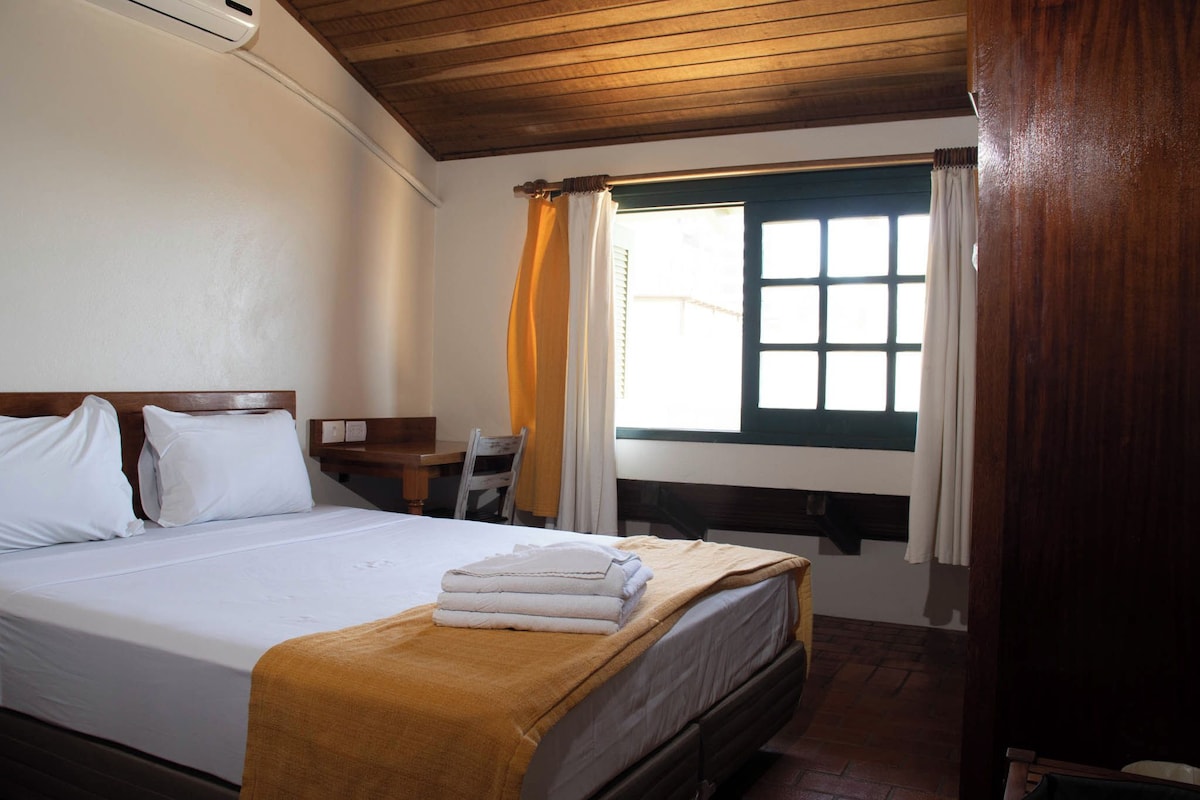 A hotel room with a bed, a desk, a window with curtains, and a wooden ceiling.