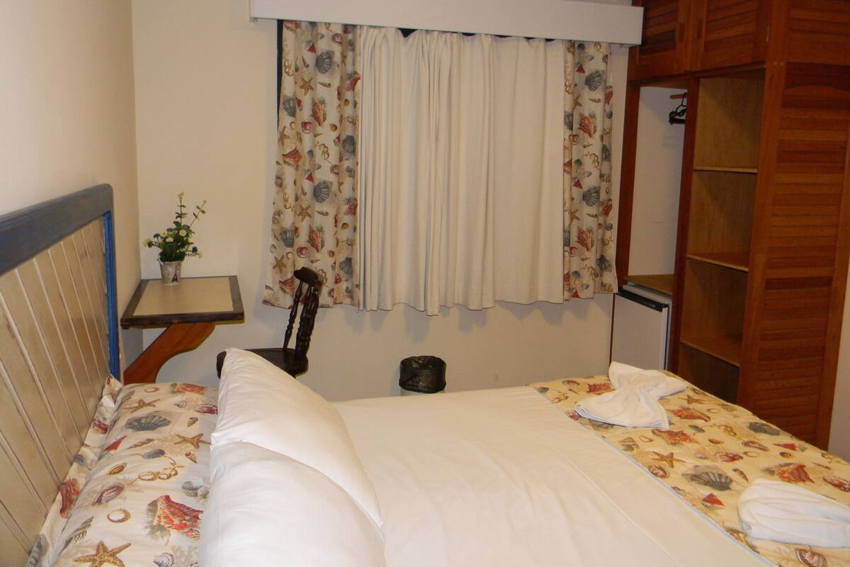 A bedroom with a bed, a small table with a plant, a guitar, and a wardrobe.