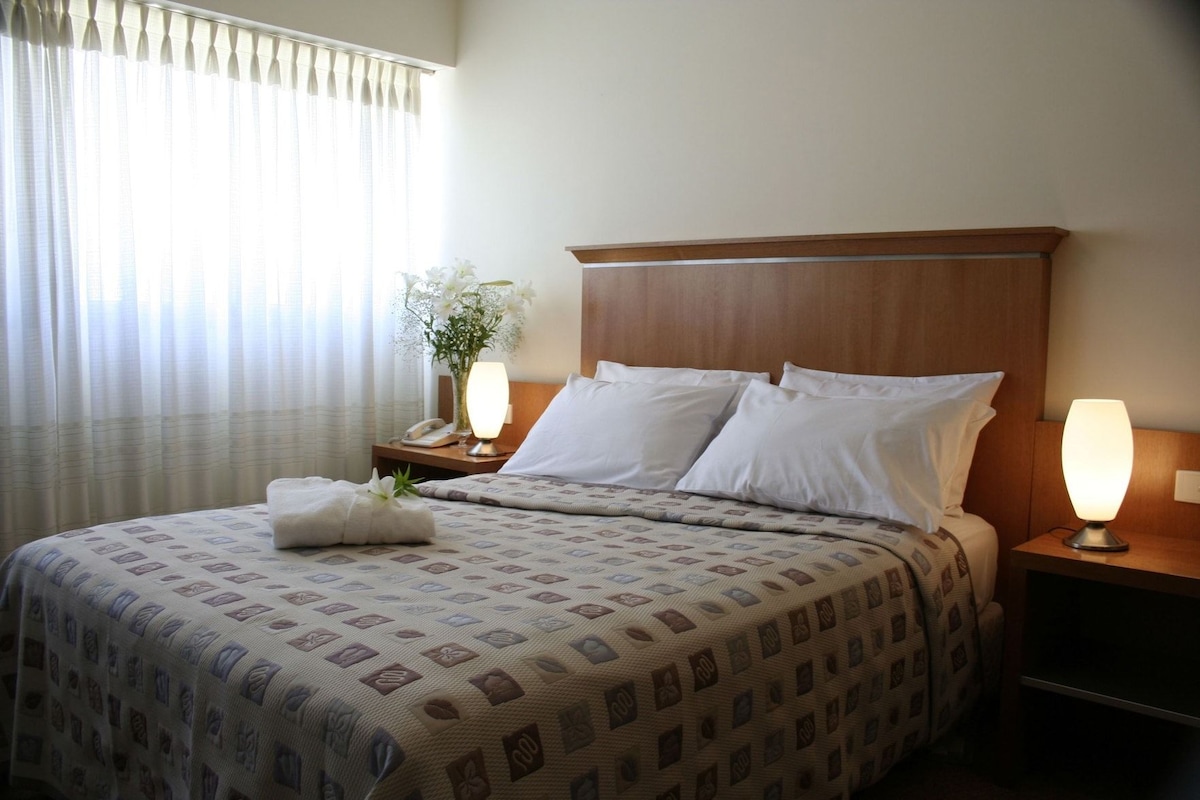 A hotel room with a bed, two bedside lamps, a nightstand with a phone, and a window with curtains.