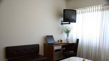 A hotel room with a bed, a desk with a laptop, a television, a chair, and a window with curtains.