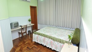 Ein Zimmer mit einem Bett, einem Schreibtisch, einem Stuhl, einem kleinen Tisch mit einer Vase und einem Fenster mit Vorhängen.