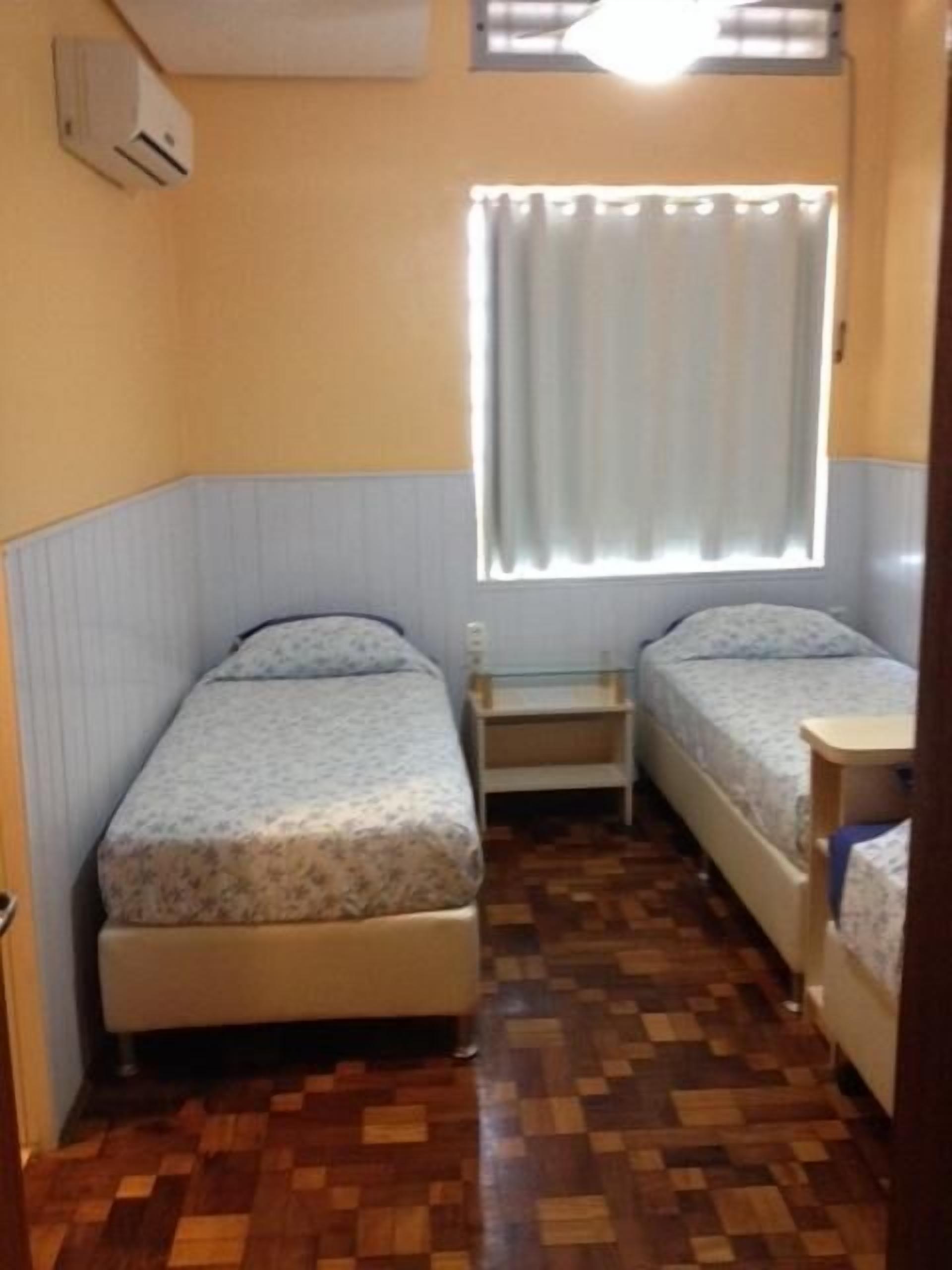 Quarto pequeno e compacto com duas camas de solteiro, uma mesinha e uma janela com cortinas.