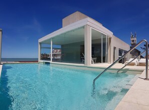 A modern poolside area with a clear view of the ocean, a covered seating area, and a lifeguard tower. - Amura Suites (Pinamar)