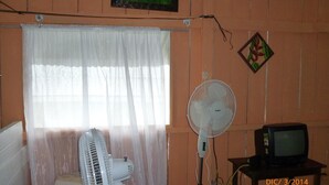 Une pièce dotée d’une grande fenêtre ornée de rideaux légers, d’un ventilateur sur pied, d’un téléviseur posé sur une table en bois et d’un tableau encadré accroché au mur.