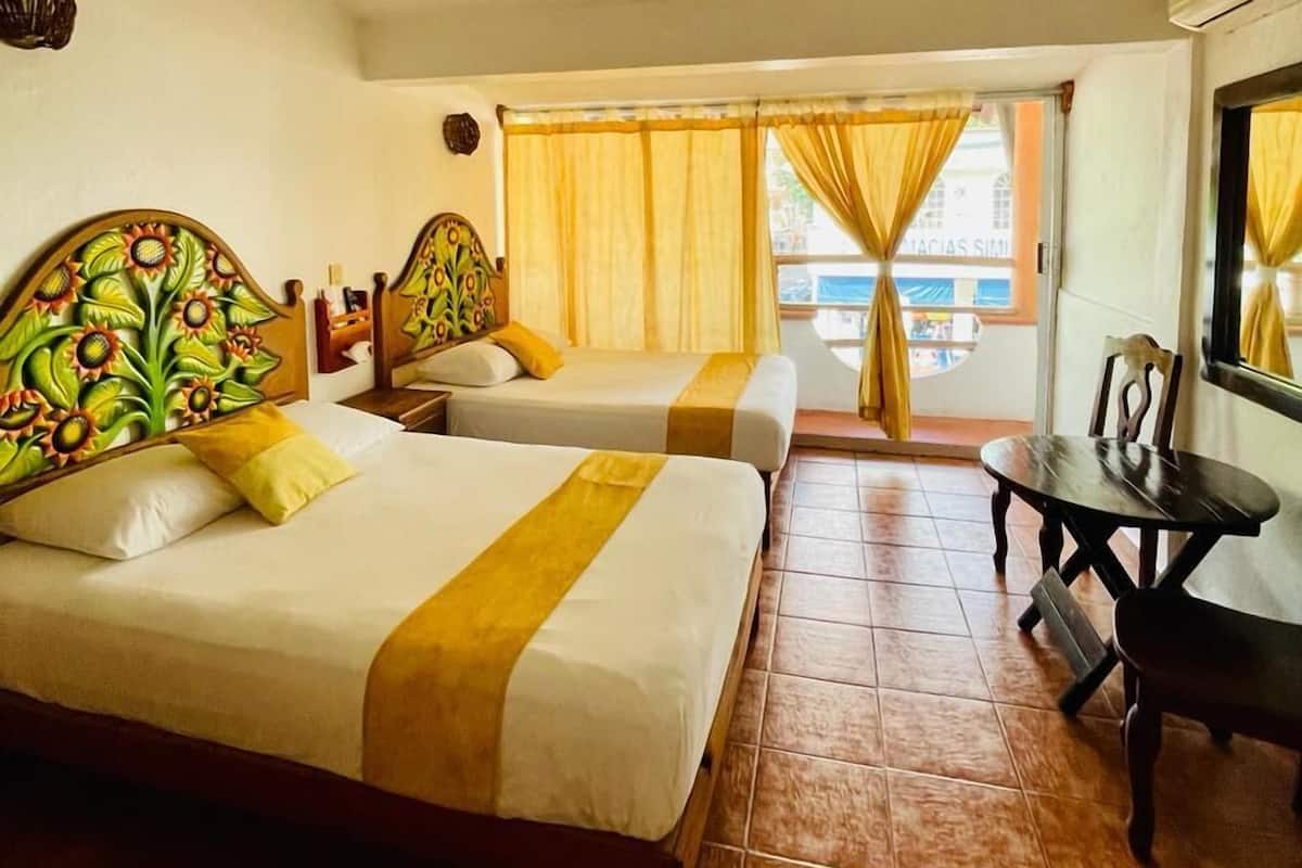 Habitación de hotel con dos camas, una mesita y vista a la calle desde la ventana.
