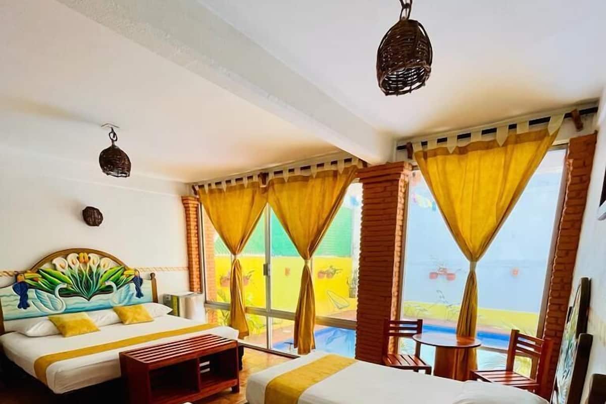 Habitación de hotel con tres camas, un banco de madera, una mesita y vistas al mar a través de amplias ventanas con cortinas amarillas.