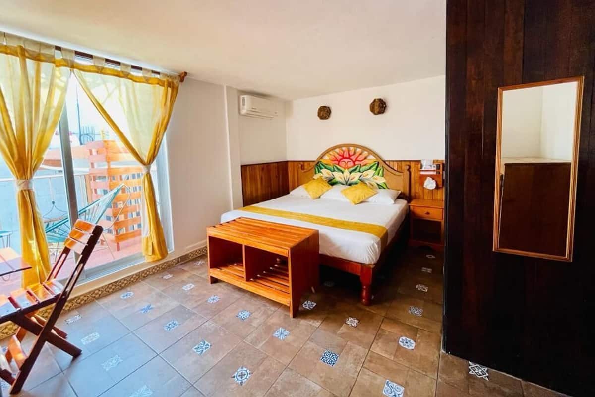 Habitación de hotel con cama, cabecera de madera, una mesita pequeña, una silla, ventana con cortinas y vistas a edificios.