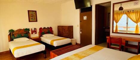Quarto de hotel com duas camas de solteiro, uma escrivaninha, uma cadeira, uma televisão e uma porta que dá para outro cômodo.