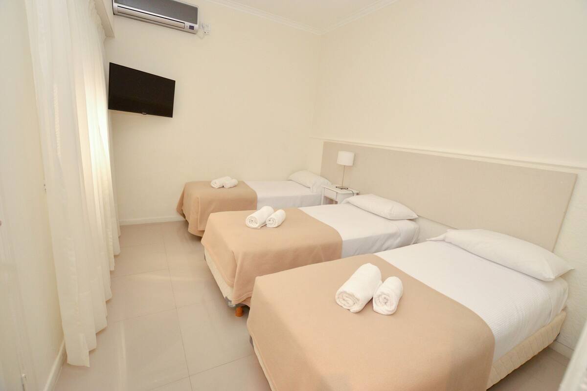 A hotel room with three single beds, each with a beige bedspread and white pillows, and a flat-screen TV mounted on the wall.