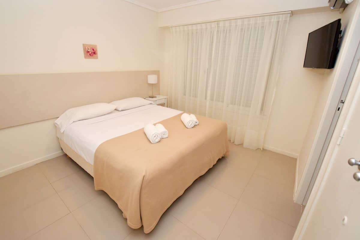 A hotel room with a bed, a nightstand, a TV, and a window with sheer curtains.