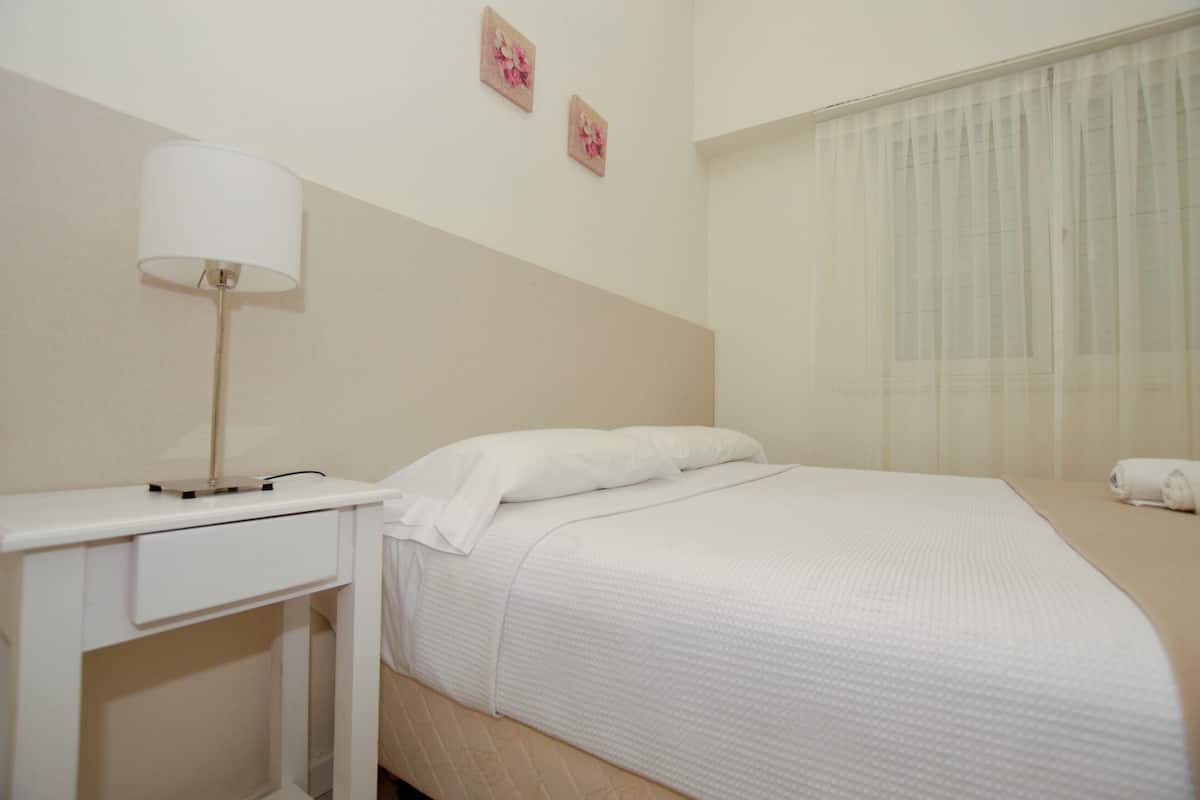 A neatly made bed with white linens, a bedside table with a lamp, and a window with sheer curtains.