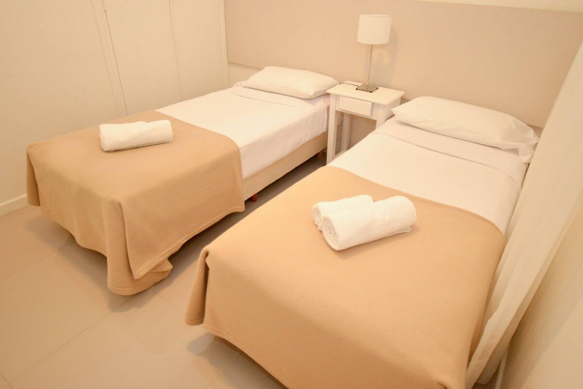 A hotel room with two beds, each with a beige bedspread and white pillows, and a small bedside table with a lamp.