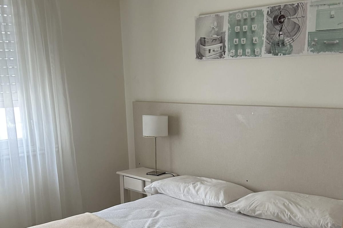 A neatly made bed with a beige blanket and rolled towels, a bedside table with a lamp, and a wall with a partial view of a calendar.