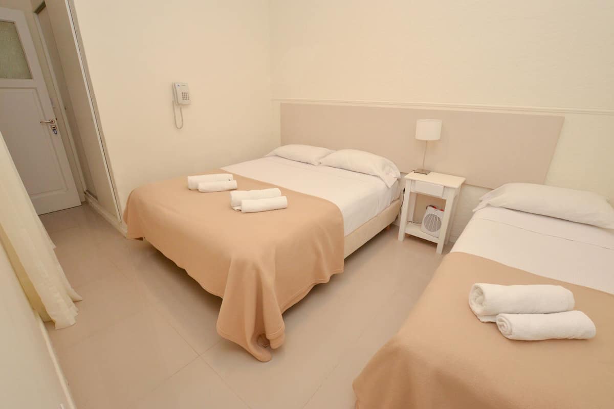 A hotel room with two beds, each with a beige bedspread and white pillows, and a bedside table with a lamp and a phone.