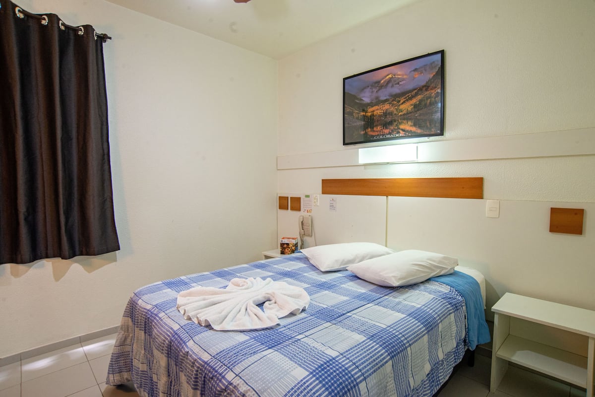 A hotel room with a bed, a nightstand, a painting on the wall, and a window with curtains.
