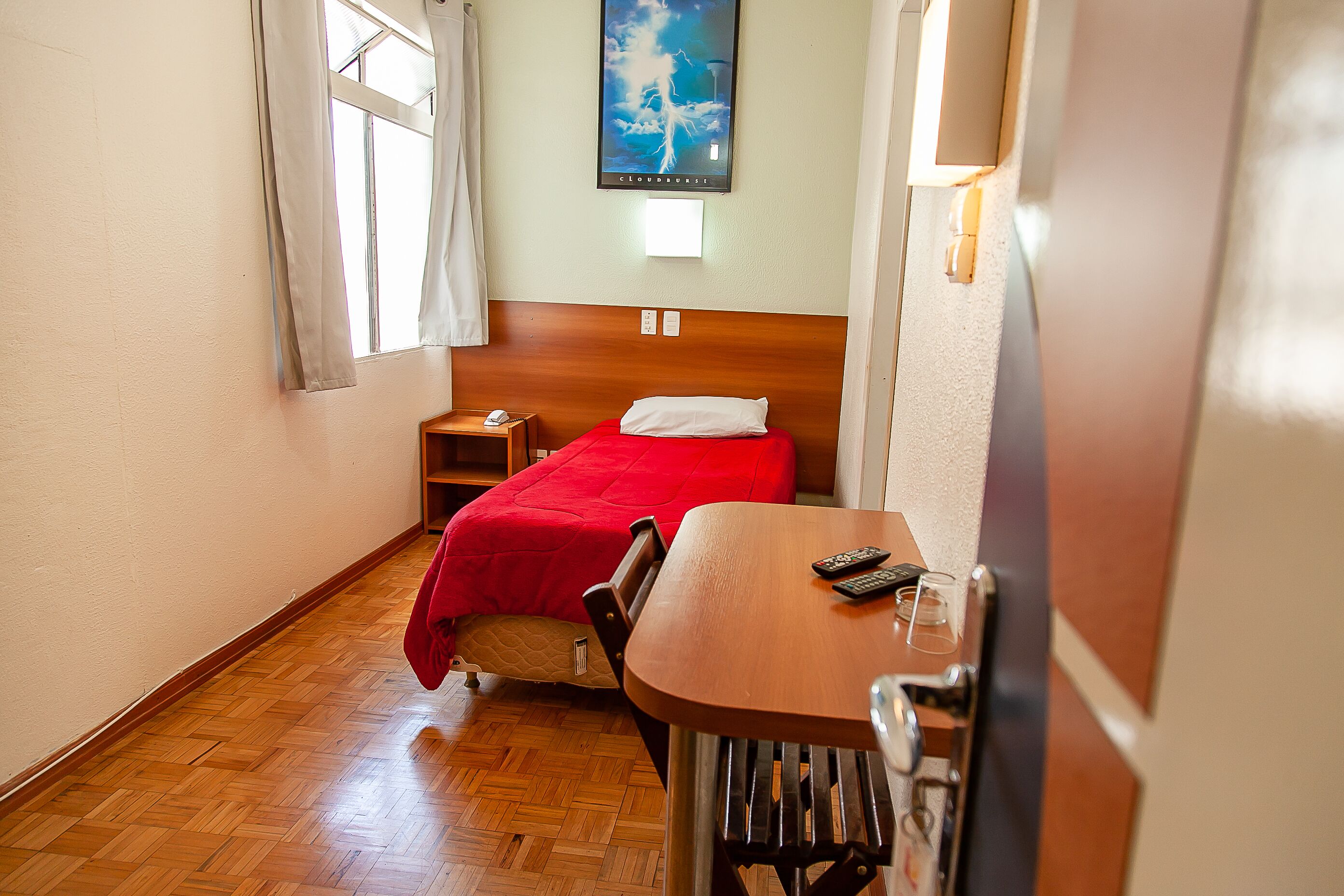 Quarto de hotel pequeno com piso de madeira, uma cama simples com manta vermelha, uma mesa de madeira com um controle remoto e uma luminária fixada na parede.