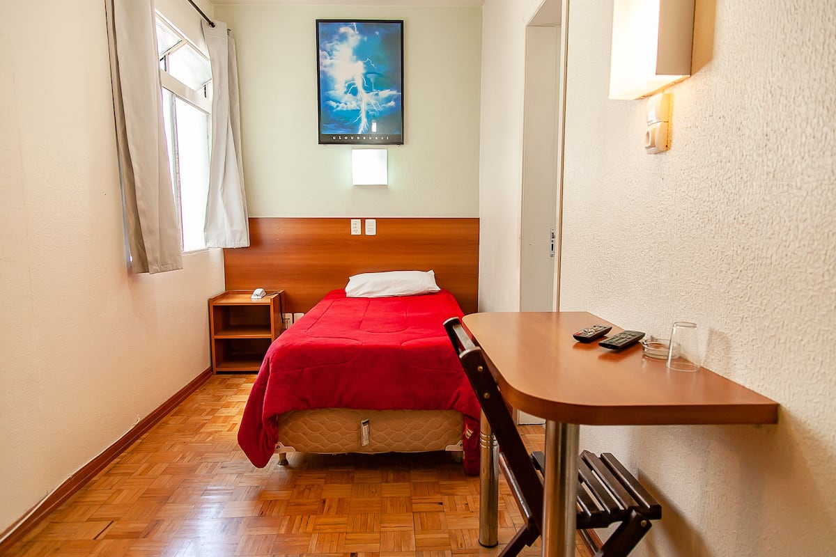 A hotel room with a wooden floor, a bed with a red blanket, a small table with a remote control, and a wall-mounted light fixture.