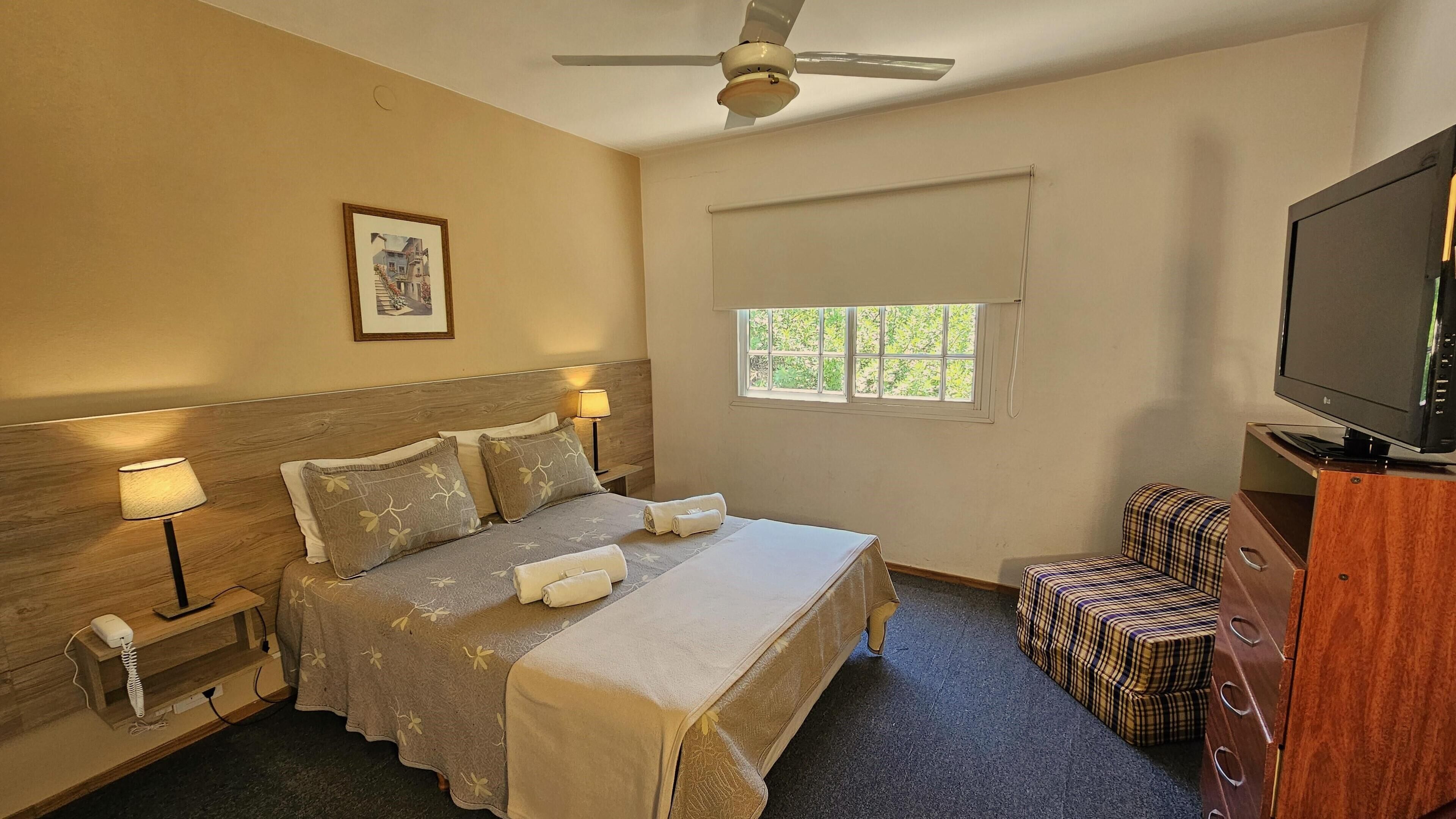 A hotel room with a bed, bedside lamps, a ceiling fan, a window with a view, a television, and a small seating area.
