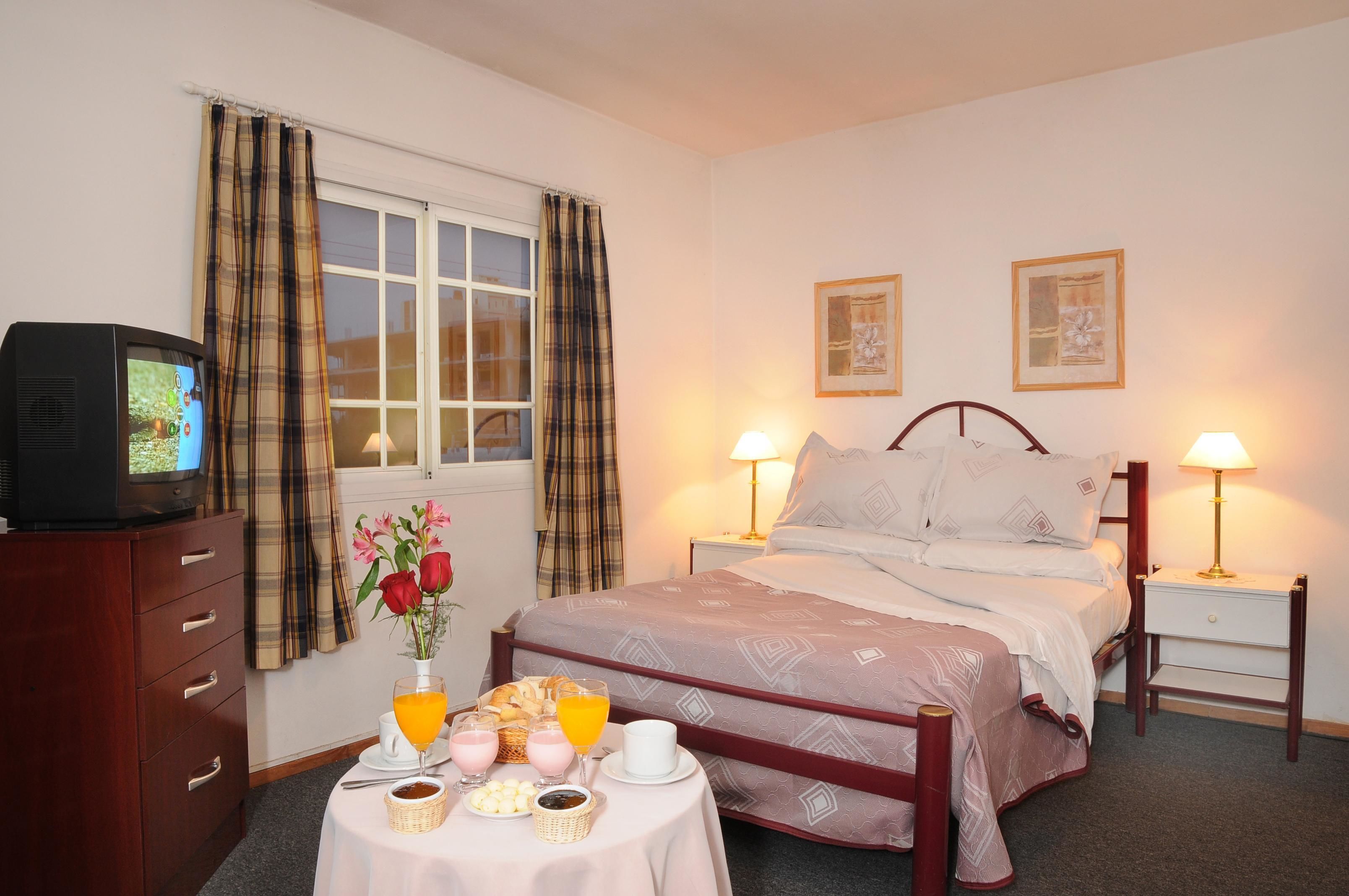 Una habitación de hotel con una cama, lámparas de noche, un televisor sobre un mueble de madera, una mesa con productos para el desayuno y dos cuadros en la pared.