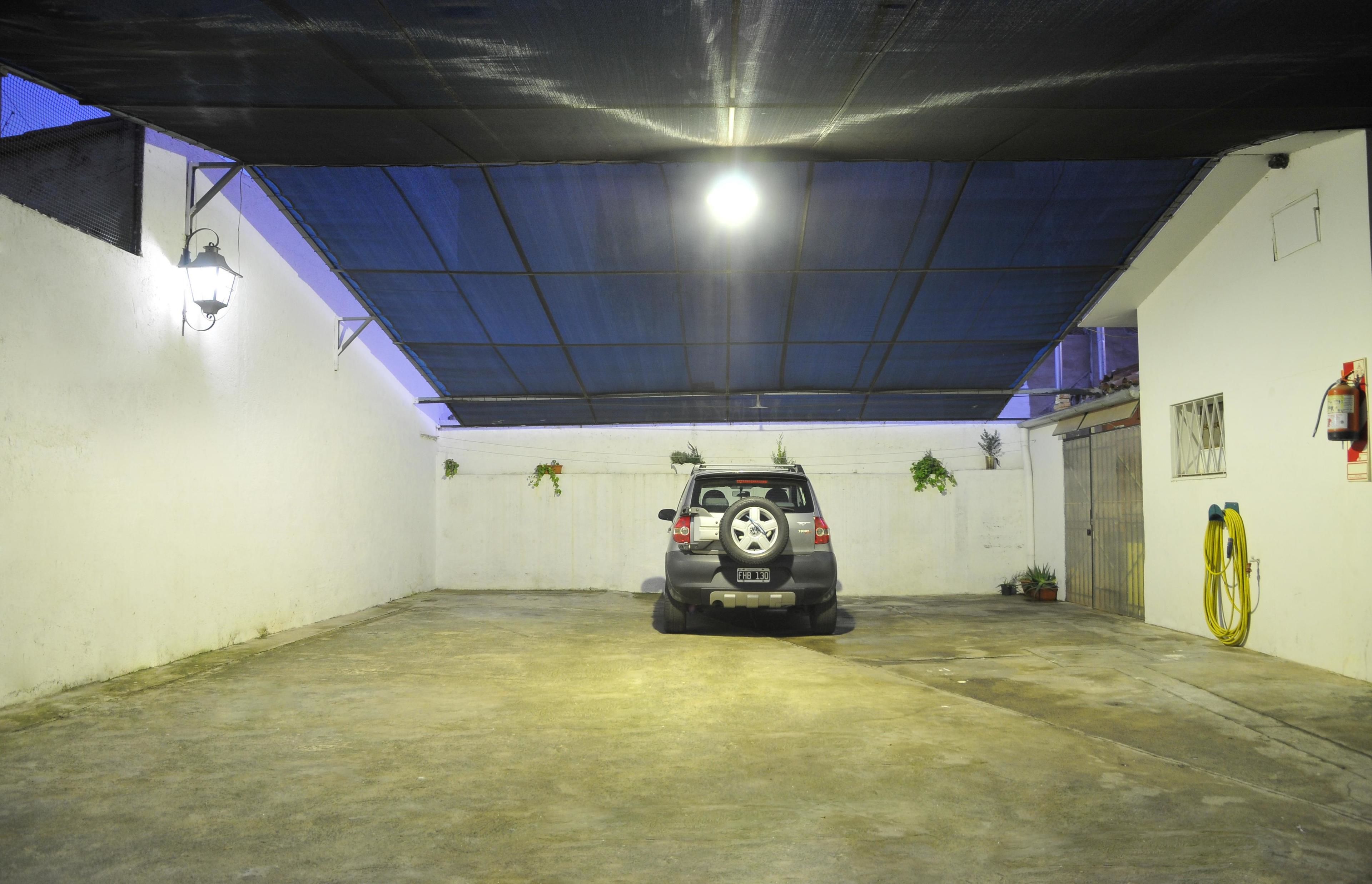 A covered parking area with a car parked in the center, a fire extinguisher on the wall, and a yellow hose coiled on the right.